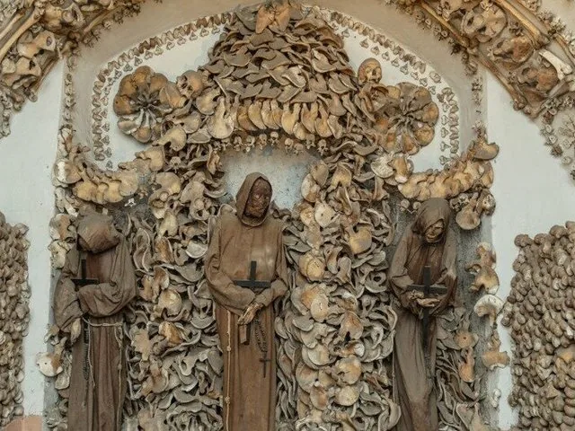 Three skeletal figures in monk habits displayed against walls elaborately decorated with human bones.