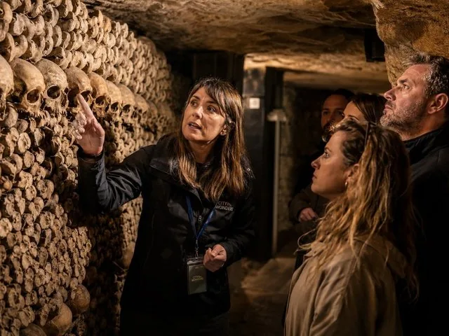 A female tour guide explaining a large wall of human skulls to a group.