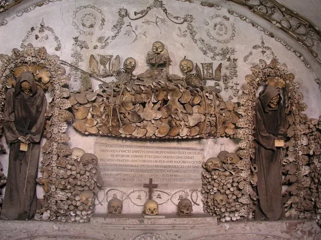 Two robed skeletons flanking an elaborate arrangement of bones and a central inscription.