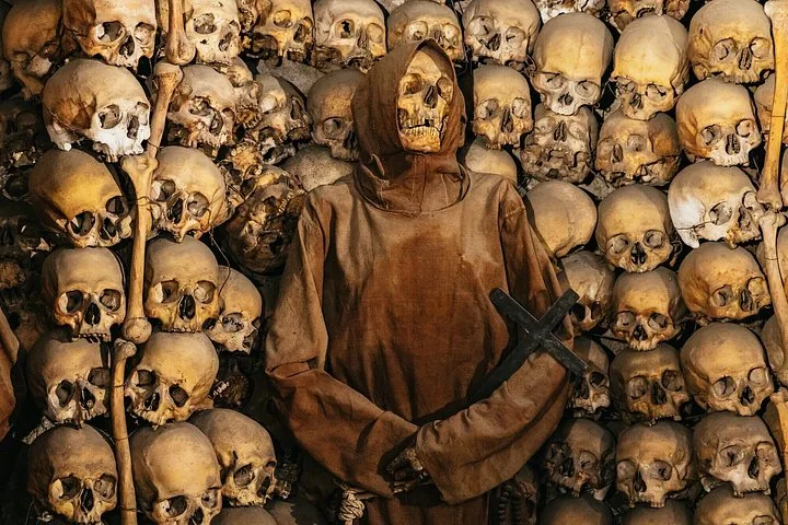 A skeleton dressed in a brown monk's habit holding a cross, surrounded by skulls.