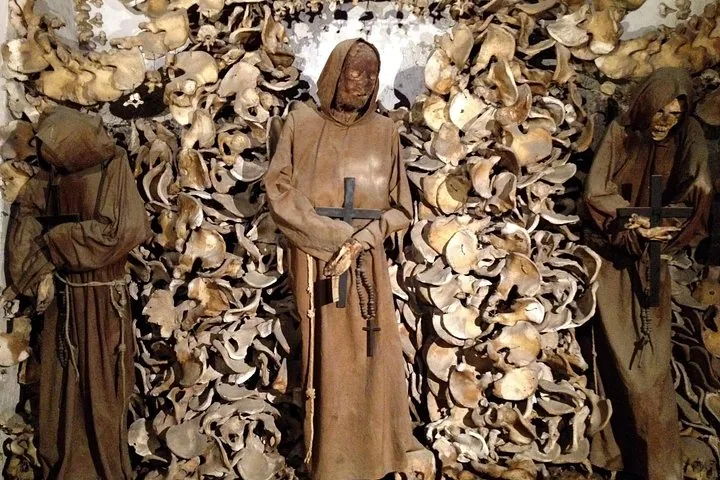 Skeletal remains dressed in monk robes holding crosses, standing against a wall of bones.
